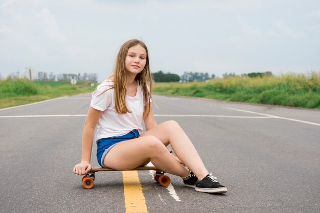 空の道でスケートボードの上に座ってモダンな魅力的なかわいい女の子