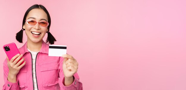 ピンクの背景の上に立っているスマートフォンでオンラインで支払う携帯電話のクレジットカードで笑って笑っている現代の美しいアジアの女の子
