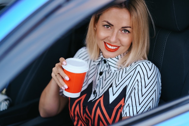 Donna bionda moderna che mangia un caffè per andare mentre si guida in città