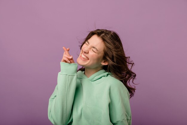 Modern caucasian young brunette woman in sweatshirt closes her eyes crosses fingers for good luck on purple background Positive emotions concept