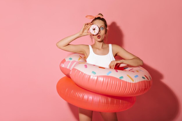 ピンクの背景にドーナツと大きな浮き輪を保持しているイヤリングと白い水着の明るいメイクでモダンなクールな女の子