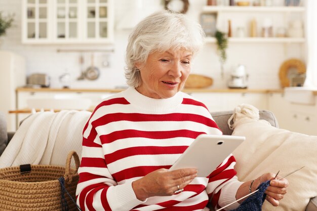 Modern electronic gadgets, devices, connection and communication concept. Attractive elderly female knitting at home, sitting on couch with yarn, watching video tutorial online via digital tablet