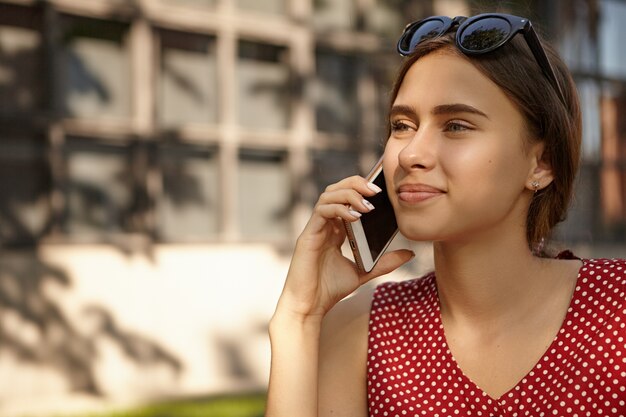 現代の電子ガジェット、人、コミュニケーションの概念。友人との素敵な会話をしながら、携帯電話で話している点線の赤いドレスを着た美しい日焼けしたヨーロッパの女性のクローズアップショット