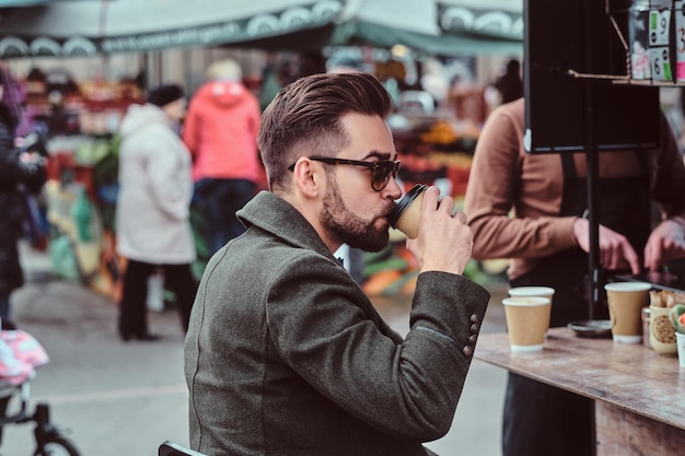 Modern elegant man in sunglasses is drinking coffee while sitting outside at coffeeshop.