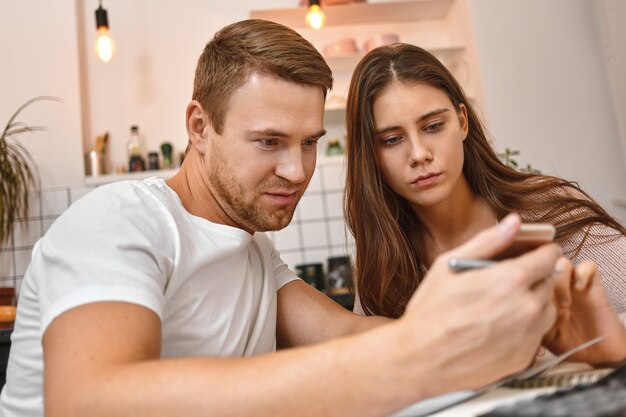 현대 가제트와 사람들. 젊은 부부는 부엌에서 커피를 마시고, 휴대 전화를 사용하여 무급 모타 주에 온라인 은행 알림을 읽고 걱정 된 표정으로 화면을보고 있습니다.