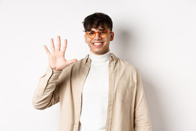 Современный парень в очках и стильном наряде, показывает номер пять пальцев и улыбается, делая заказ, стоя на белом фоне
