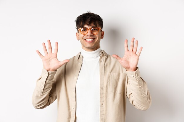 Modern guy in glasses and stylish outfit, showing ten fingers number and smiling, standing on white background.