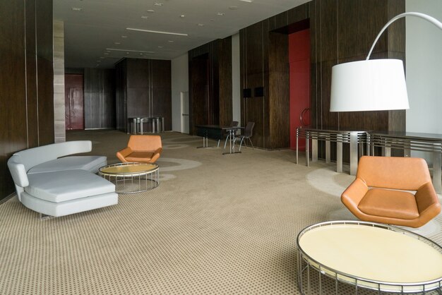 Современный вестибюль отеля с кожаным диваном и стульями, лампой и круглыми столами.