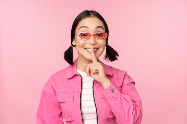 Modern korean girl in stylish spring outfit sunglasses showing shush hush sign press finger to lips taboo gesture standing over pink background