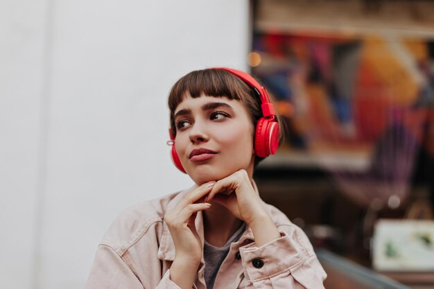 Modern lady with red headphones posing outside Shorthaired girl in denim cool clothes looking away and posing outdoors