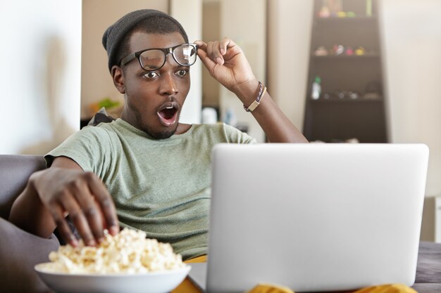 現代のライフスタイル、技術、人々の概念。驚いた若いアフリカ系アメリカ人男性は、仕事の後に自宅でリラックスしてバスケットボールの試合をオンラインで見たり、ソーシャルメディアのビデオを見たり、ポップコーンを食べたりしています