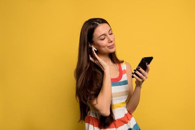 Modern lovely woman with long dark hair wearing brightful dress listening music with smartphone over yellow wall.