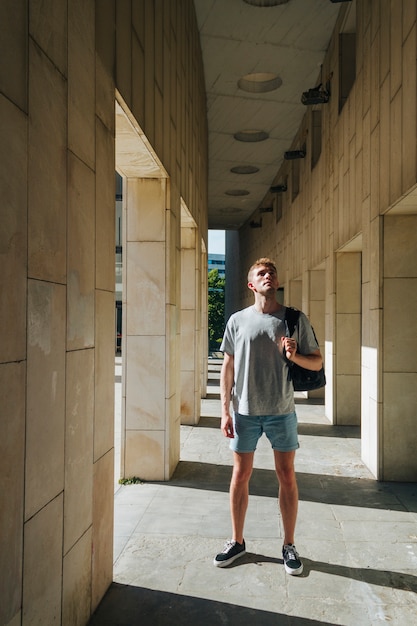 Modern man carrying backpack standing near pillar and looking up
