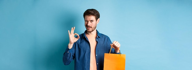 Foto gratuita uomo moderno in abito casual che mostra la borsa della spesa e il segno ok che fa l'occhiolino alla telecamera raccomandando il negozio s