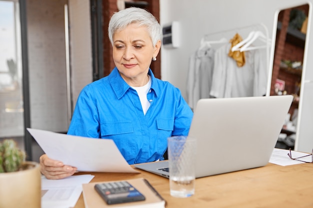 Modern middle aged woman with stylish short hair reading piece of paper in her hand working remotely on generic laptop pc, sitting at desk with calculator and copybook in cozy home interior