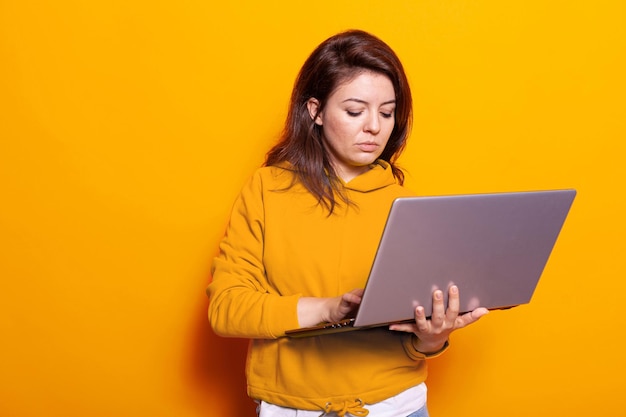 Modern person holding laptop to use technology on camera in studio. Portrait of young woman looking at digital device screen and working online while standing over isolated background.