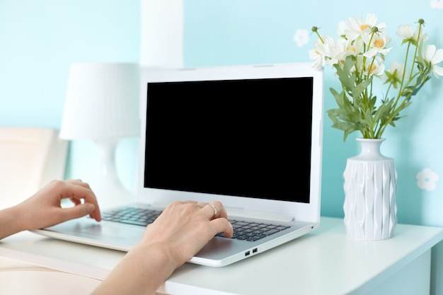 Modern portable laptop computer with blank screen on desk surrounded with lamp and bouquet on blue wall. Unrecognizable woman works distantly on modern electonic device, connected to wifi