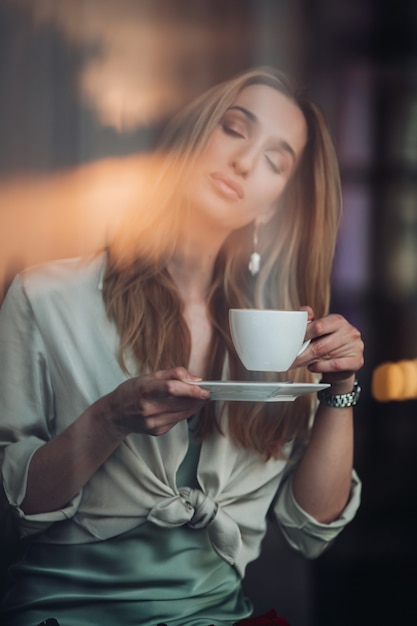Бесплатное фото Современная романтическая красивая молодая девушка с закрытыми глазами наслаждается ароматом кофейной чашки во время отдыха в кафе в помещении