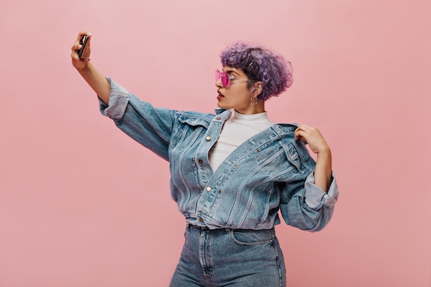 Modern short-haired woman with a bright hairstyle in pink glasses makes selfie. Beautiful curly woman in denim jacket posing.