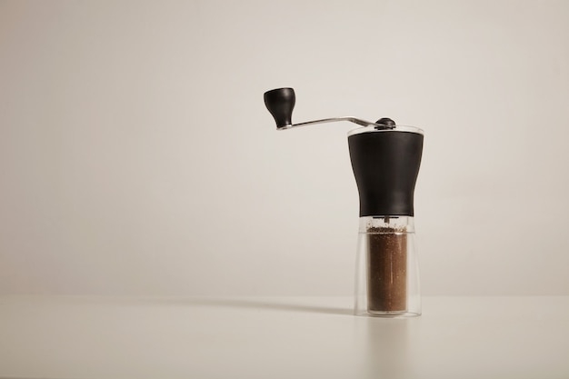 Modern slim manual burr grinder with freshly ground coffee on white table on white wall