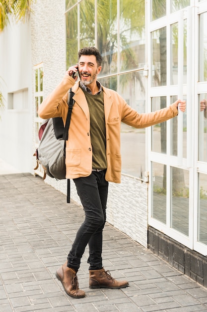 Modern smiling man with his backpack talking on mobile phone while opening the glass door