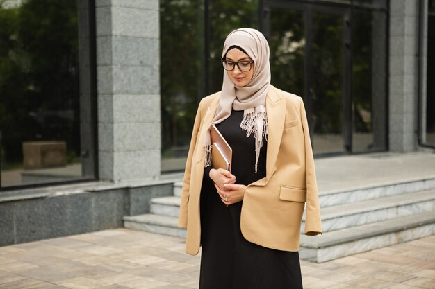 Modern stylish muslim woman in hijab, business style jacket and black abaya walking in city street with laptop