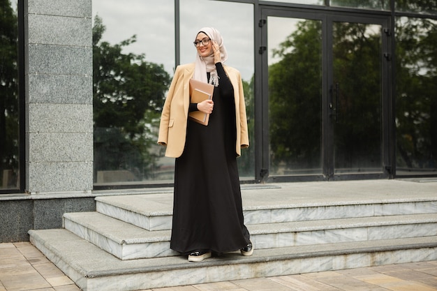 ヒジャーブを着たモダンでスタイリッシュなイスラム教徒の女性、ビジネス スタイルのジャケット、ノート パソコンを持って街を歩く黒いアバヤ