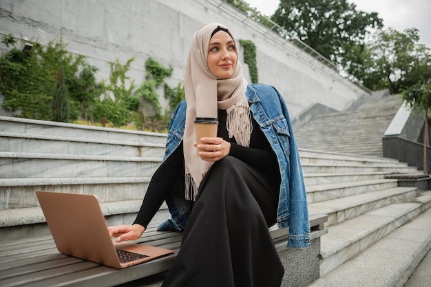 Modern stylish muslim woman in hijab, denim jacket and black abaya sitting in city street working on laptop