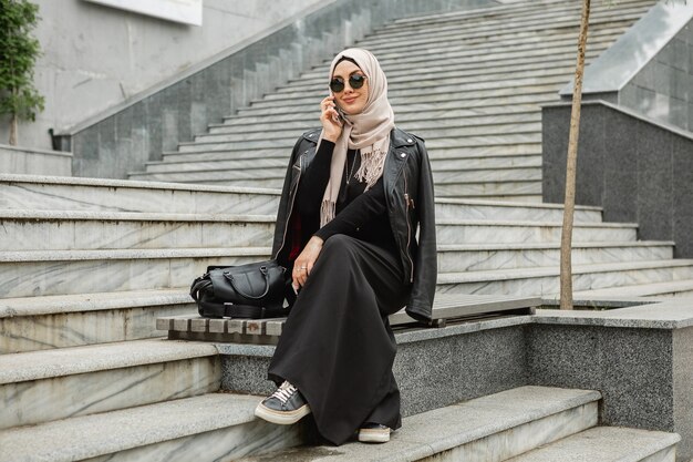 hijab, 가죽 재킷 및 스마트 폰에 얘기하는 도시 거리에서 걷는 검은 아바야의 현대적인 세련 된 이슬람 여성
