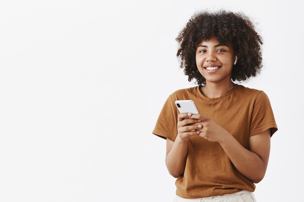 modern stylish and pleased african american woman using wireless earphones while talking with clients holding smartphone making notes or picking music in device