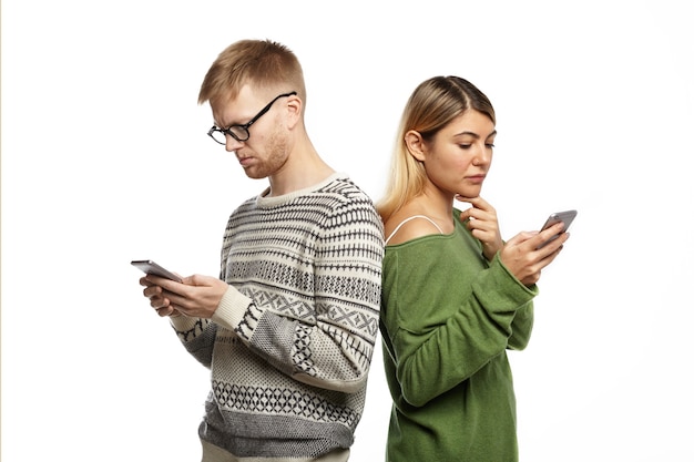 Современные технологии и концепция интернет-зависимости. Стильная молодая пара мужчина и женщина стоят спиной к спине, поглощенные электронными гаджетами, не глядя и не разговаривая друг с другом, обмениваясь сообщениями онлайн