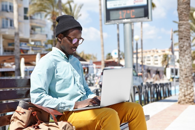 현대 기술, 사람 및 도시 생활. 매력적인 젊은 아프리카 계 미국인 블로거 리조트 타운에서 휴가 기간 동안 일반 노트북 PC를 사용하여 새 기사를 작성하고 외모에 영감을주었습니다.