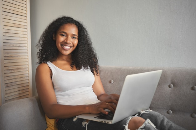 현대 기술, 통신 및 전자 기기 개념. 그녀의 무릎에 휴대용 컴퓨터와 함께 소파에 앉아 먼 거리에서 일하는 아름다운 긍정적 인 젊은 어두운 피부 여자 카피라이터의 그림