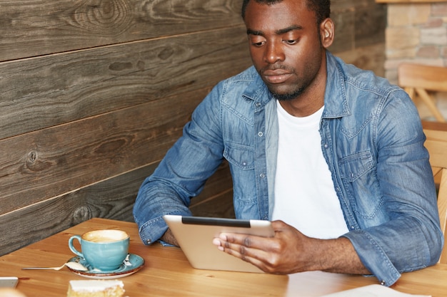 Tecnologia e comunicazione moderne. elegante studente africano che naviga in internet su tavoletta digitale, godendo della connessione wireless gratuita al bar durante la pausa caffè. messaggistica online maschile dalla pelle scura