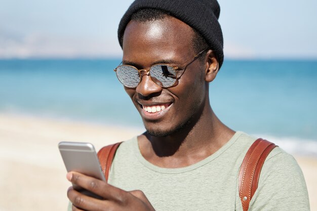 Modern technology, lifestyle, travel and tourism. Happy Afro American male traveler typing text message on smartphone, looking at screen with broad smile during walk at seaside on sunny summer day