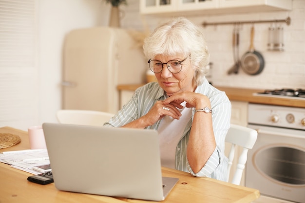 현대 기술, 노인 및 은퇴. 온라인 대출 신청서를 작성하는 안경에 회색 머리 여성 연금 수령자, 노트북 앞에 앉아 진지하게 집중된 표정으로 정보 읽기