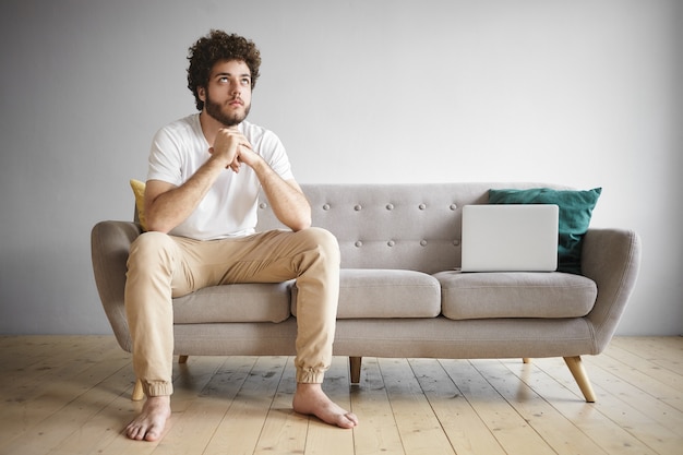 Modern technology, people and lifestyle concept. Portrait of attractive young unshaven man with voluminous hair and bare feet having tired expression, looking up while working on laptop on couch