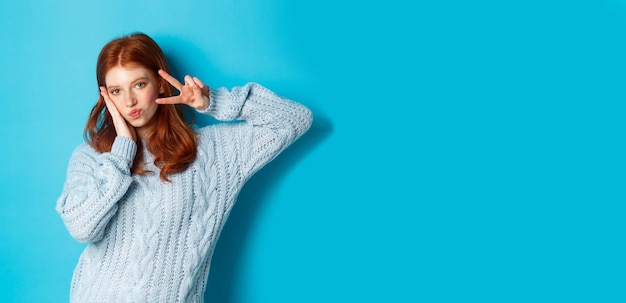 Бесплатное фото Современная девушка-подросток с рыжими волосами показывает знак мира и позирует в свитере на синем фоне