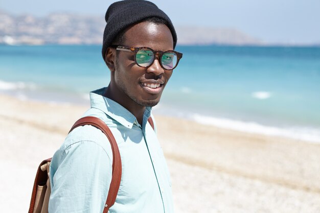 Современный модно выглядящий молодой черный европейский мужской турист в очках и рюкзаке с шляпой, проводящим солнечный весенний день на городском пляже, радуясь хорошей погоде и спокойному морю путешествия и туризм