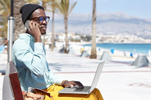 Современный модно выглядящий молодой темнокожий бизнесмен, работающий удаленно на ноутбуке и использующий мобильный телефон для совершения деловых звонков, сидя на набережной вдоль берега синего моря в солнечный день