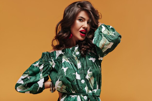 Modern wavy haired brunette with red earrings in green and white printed dress looking into camera on isolated orange backdrop