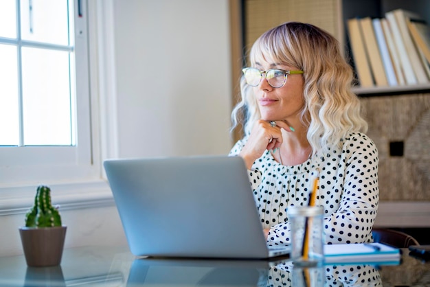 現代のウェブワーカーの大人の女性は、窓の外を見て考えているホームオフィスのワークステーションのデスクトップで休憩してリラックスした時間を過ごしています。ラップトップと接続で幸せなビジネスマン
