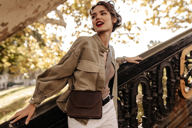 Modern woman in denim jacket and white pants looking away outside. Wavy-haired woman with red lips with handbag posing near stairs.