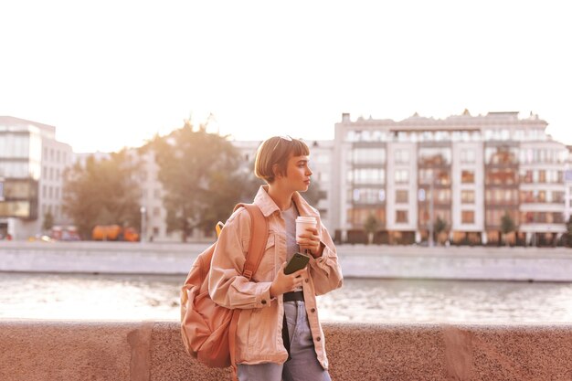 Modern woman in denim outfit with brown backpack holding cup of coffee in city