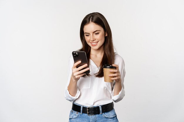 휴대 전화를 보고 흰색 배경 위에 서 있는 테이크아웃 컵 커피를 마시는 현대 여성