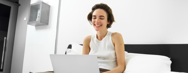 Modern woman resting at home sitting on bed with laptop looking at computer screen laughing and