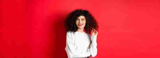 Free photo modern woman in sweatshirt playing with curly hair and looking aside at empty space standing on red