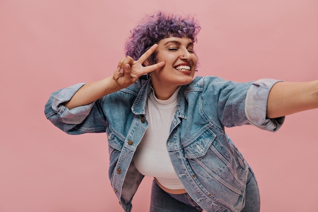 Foto gratuita la donna moderna con i capelli viola ricci luminosi mostra il segno di pace. la signora sorridente con il piercing al naso prende selfie.