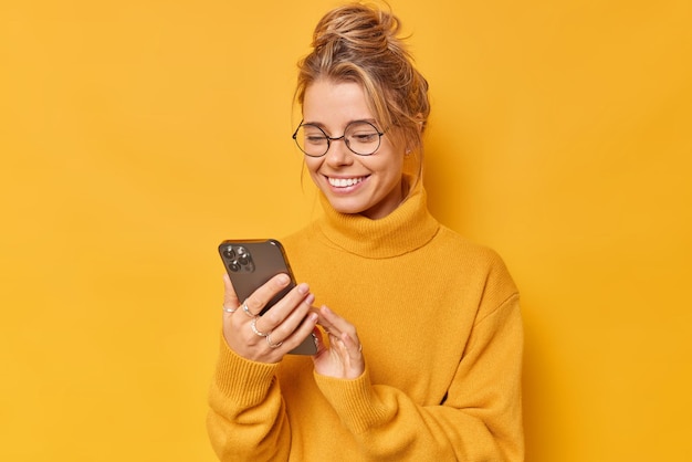 빗질된 머리를 한 현대 여성은 노란색 배경 다운로드 뉴스 응용 프로그램에 격리된 둥근 안경 캐주얼 점퍼를 착용하고 즐거운 메시지를 받는 것을 기쁘게 생각하는 휴대 전화를 읽거나 채팅을 합니다.