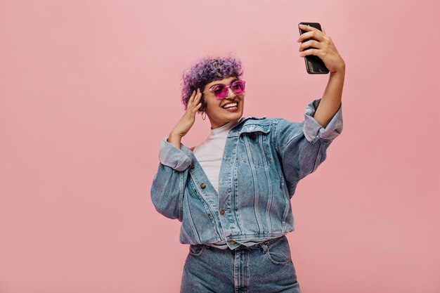 Modern woman with purple hair in cool sunglasses makes photo on pink. Smiling lady in wide bright clothes takes selfie.